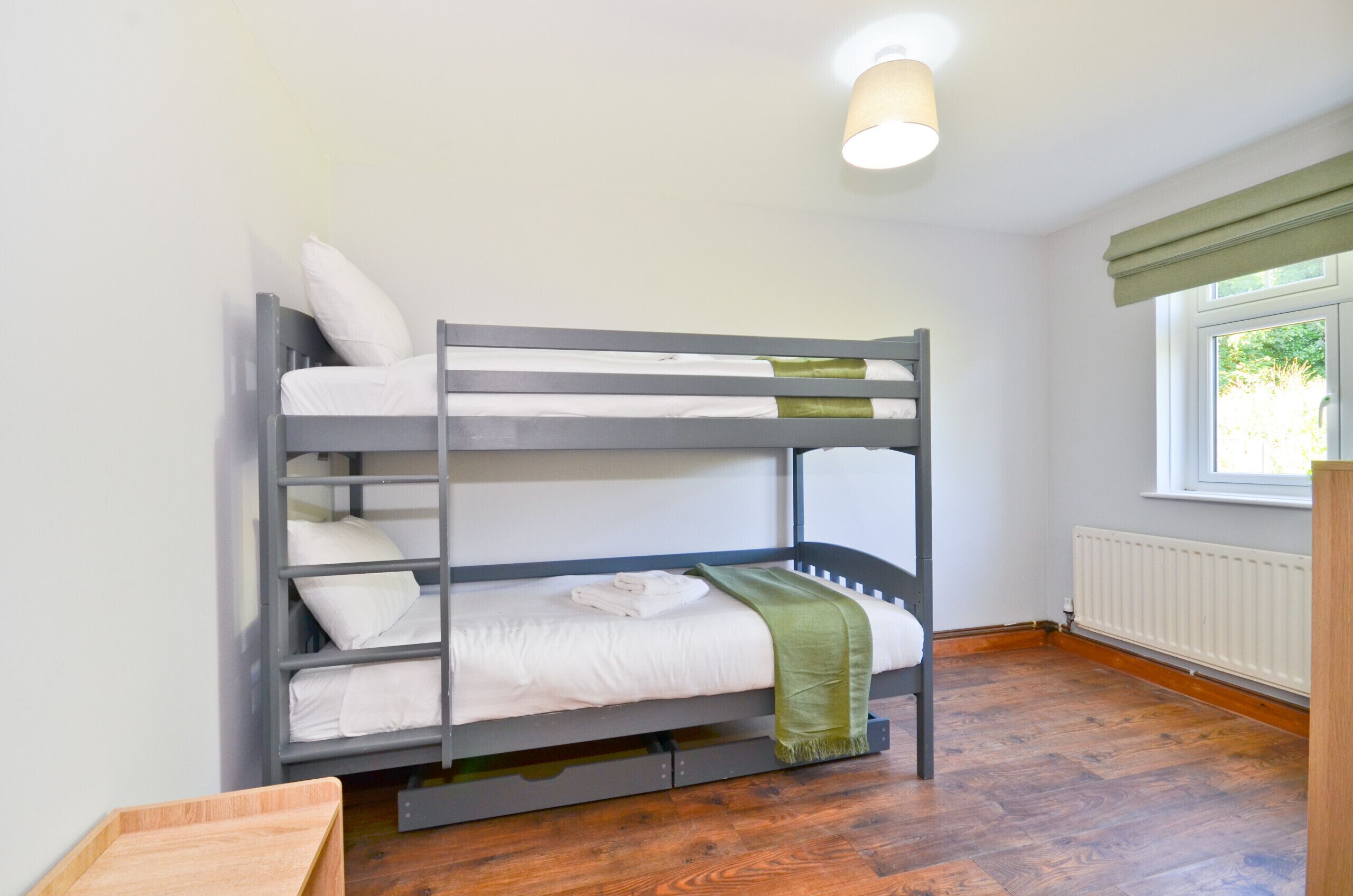 Un letto a castello con biancheria bianca e una coperta verde, pavimento in legno e una finestra vista sul verde.