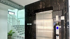En hiss med en digital display och knappar för att underlätta användningen för personer med funktionsnedsättning, en trappa med en krukväxt och en modern inredning med marmorgolv och svarta väggar med guldfärgade inslag.