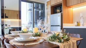 A dining area with a wooden table set for a meal, a kitchen in the background, and a view through a glass door. - Lakeside Residences by NLA (Choeng Thale)