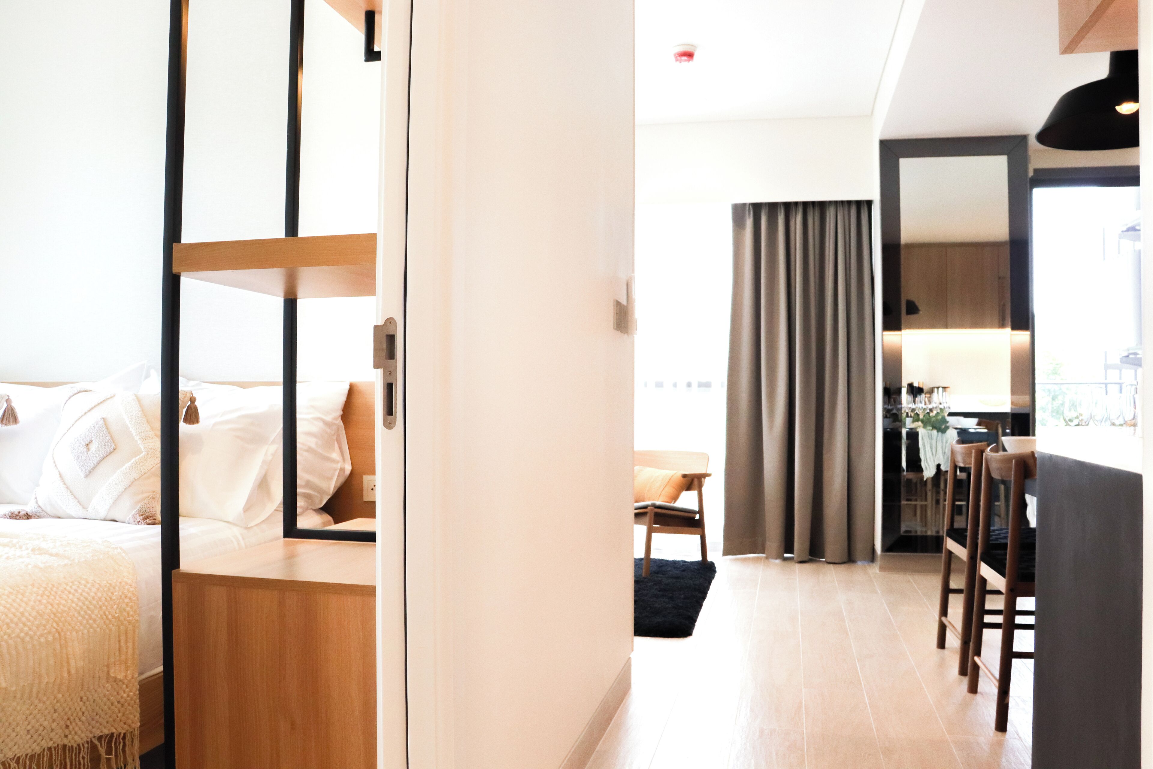 A modern hotel room with a bed, a wooden nightstand, a desk, and a dining area with chairs and a table.