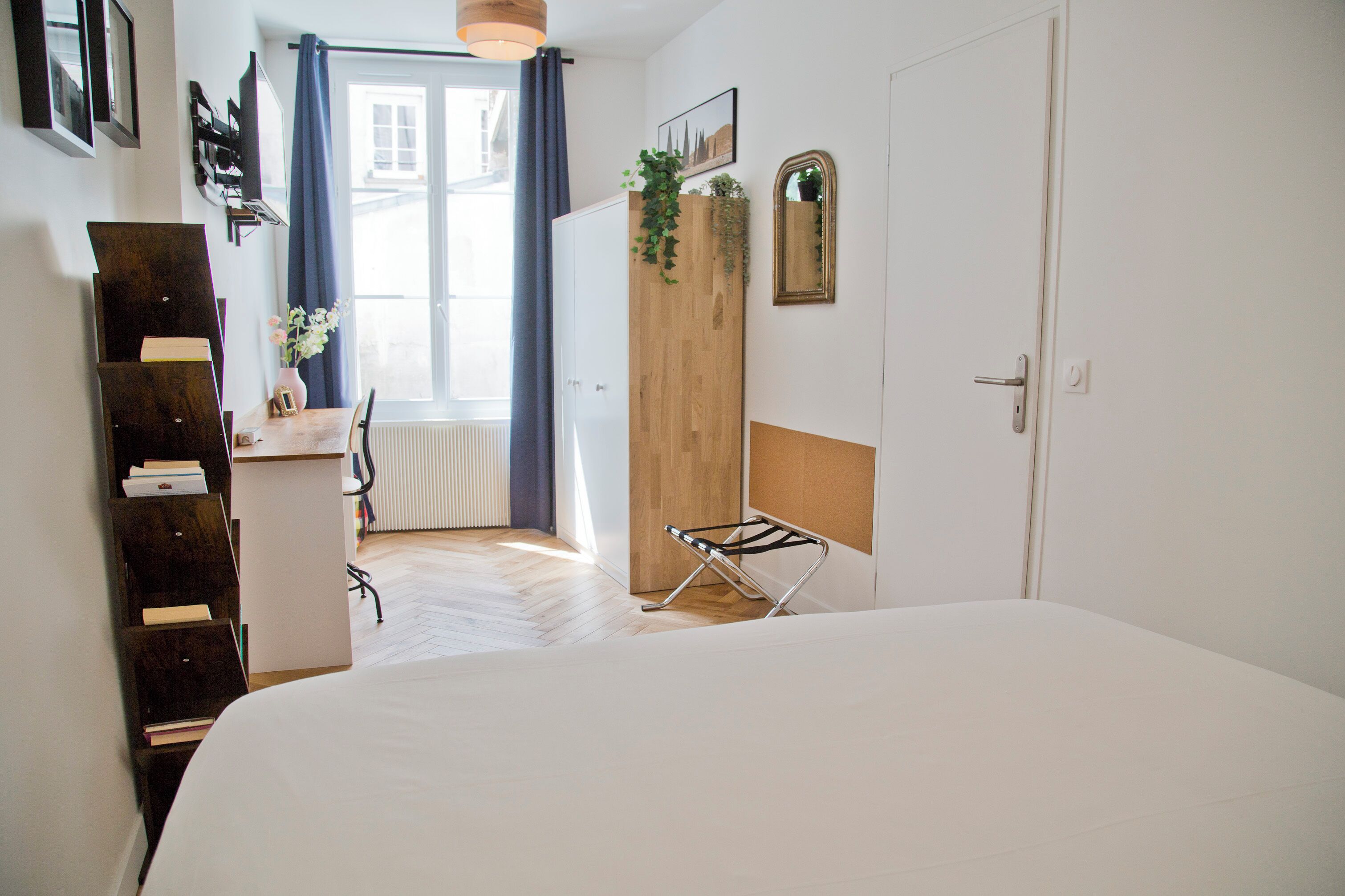 Une chambre avec un grand lit, une armoire en bois, un radiateur, une fenêtre avec des rideaux et une petite table sur laquelle est posé un vase de fleurs.