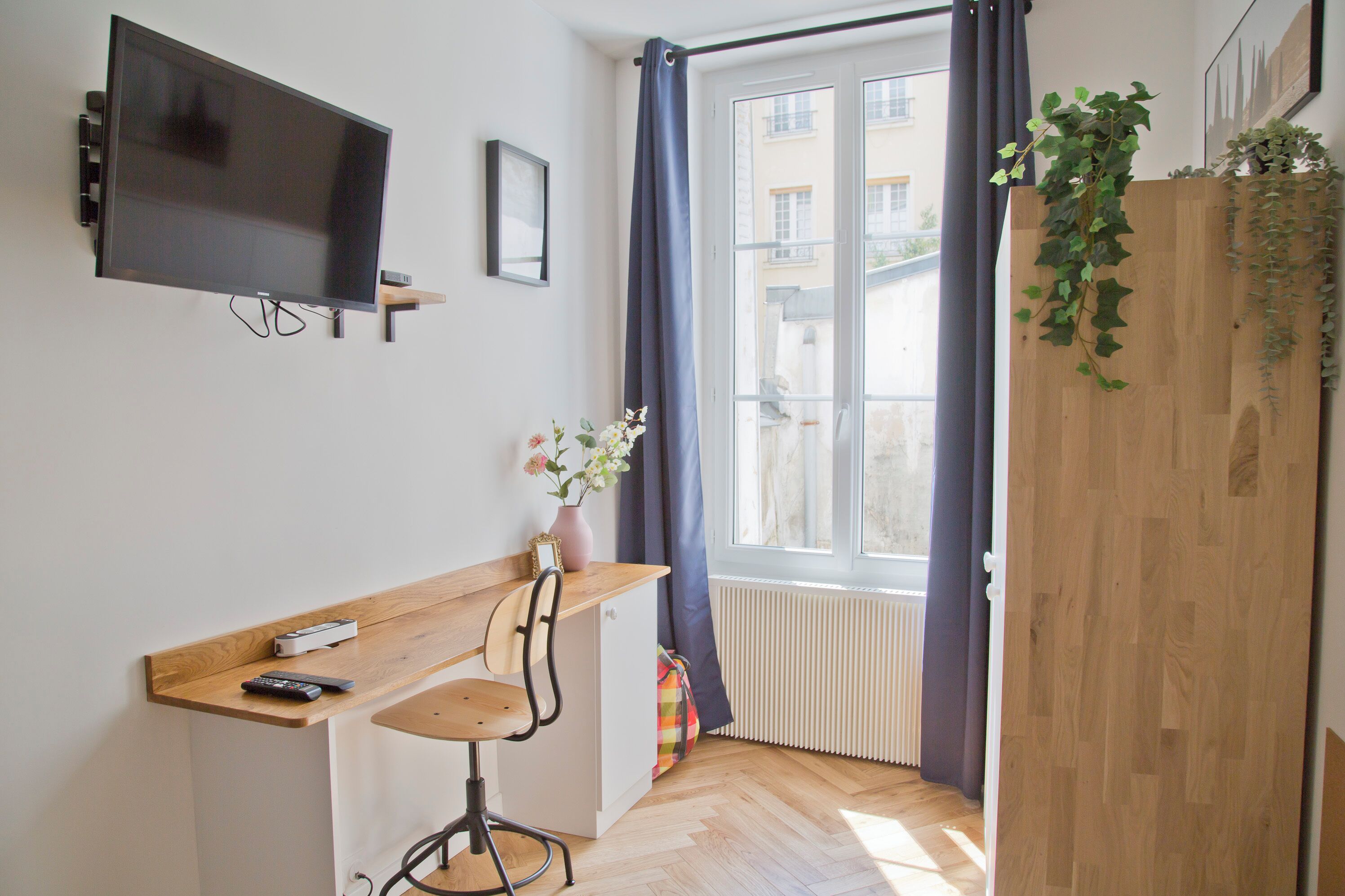 Een kamer met een aan de muur bevestigde televisie, een houten bureau met een stoel, een radiator en een raam met gordijnen.