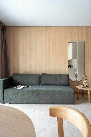 A modern living room with a grey sofa, a round wooden coffee table, a small side table with a decorative sculpture, and a large mirror on the wall.