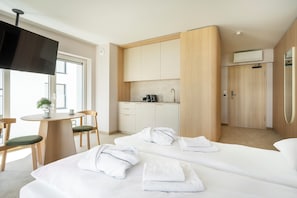 A hotel room with a bed, a desk, a chair, a small table, a plant, and a kitchenette.