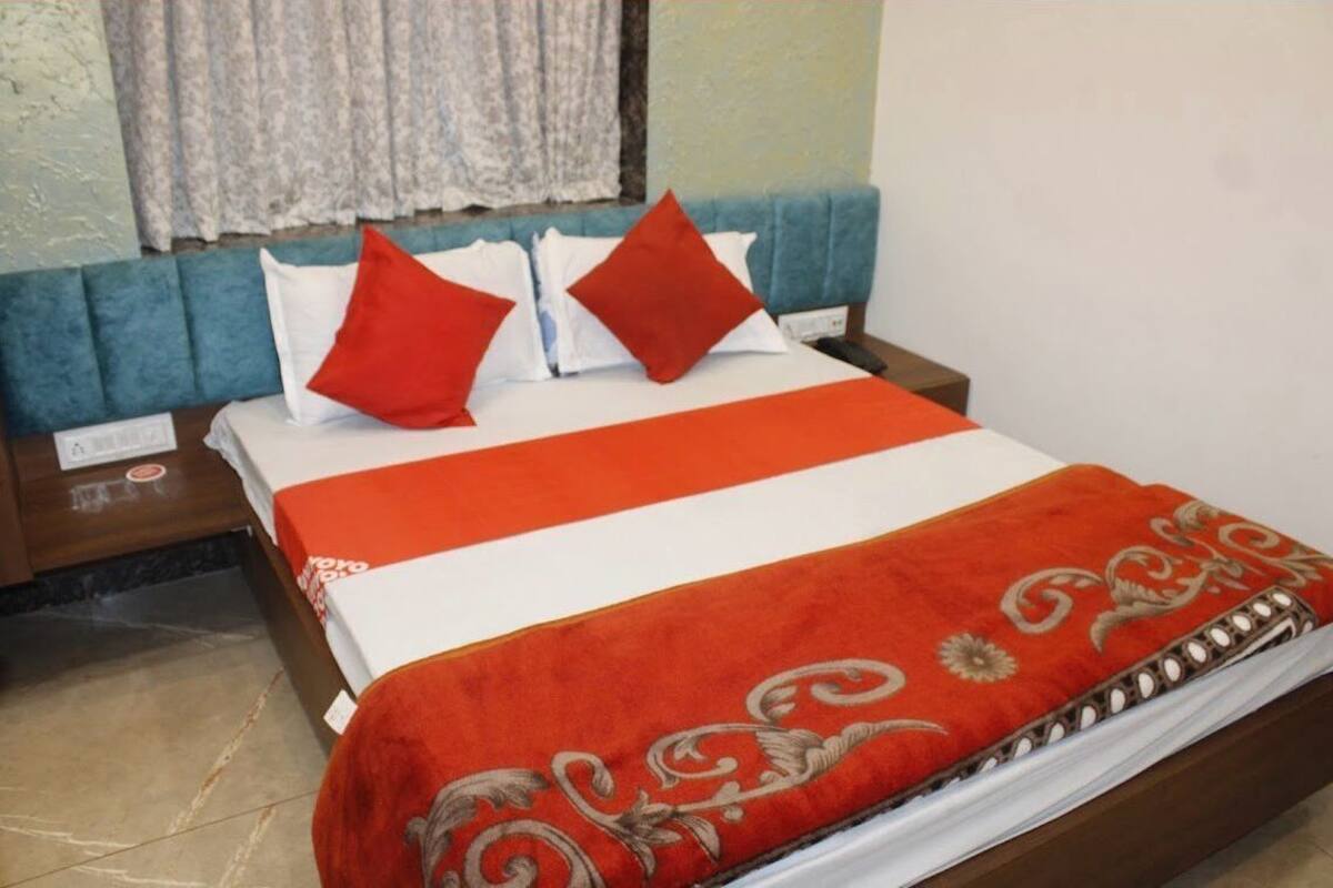 Un lit bien fait, recouvert d’une couvre-lit rouge et blanche, avec deux oreillers rouges, une tête de lit en bois et une table de chevet surmontée d’une lampe.