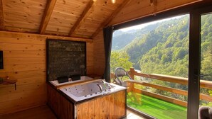 A wooden cabin with a hot tub, a stone fireplace, and a balcony with a view of the forest.