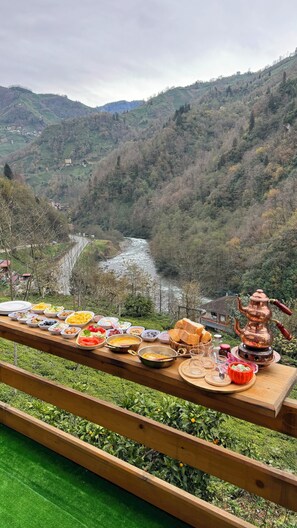 Une table à manger dressée avec différents plats et une théière en cuivre, le tout mis en valeur par un paysage de rivière et de montagnes en arrière-plan.
