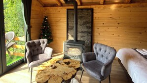 A cozy cabin interior with a wooden ceiling, a fireplace, two armchairs, a round coffee table, and a bed.