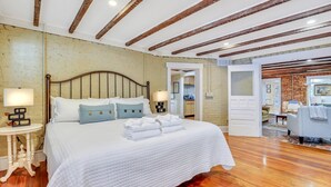 A bedroom with a large bed, wooden beams on the ceiling, and a view into the living area.