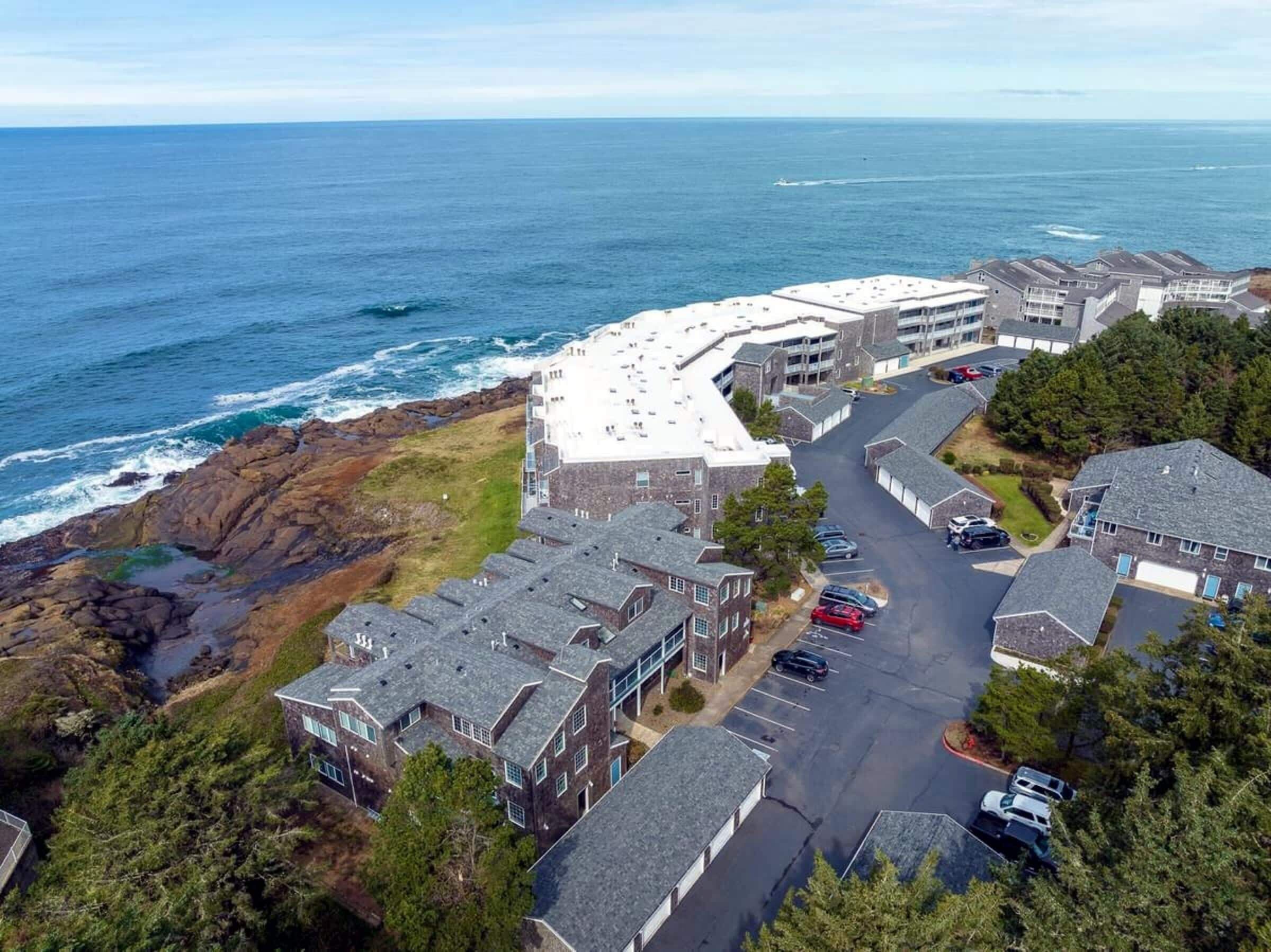 A coastal residential area with a mix of modern and traditional architecture, featuring a prominent white building with a flat roof and a parking lot with several cars.