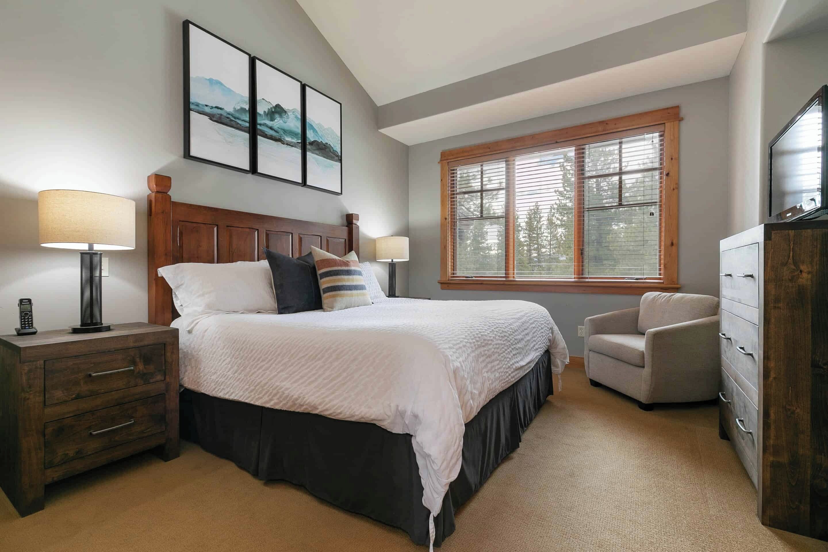 A bedroom with a bed, bedside lamps, a nightstand, a chair, a dresser, and a window with blinds.
