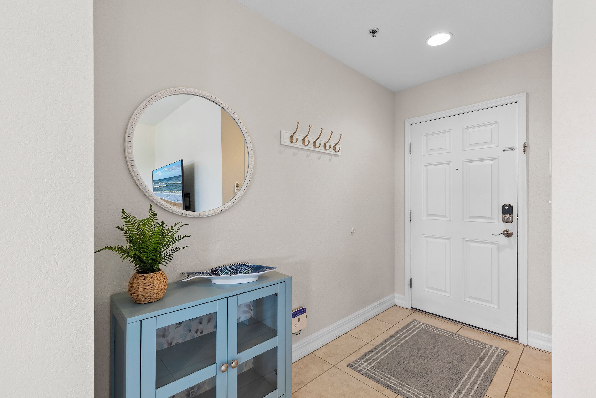 A hallway with a white door, a blue cabinet with a plant, a mirror, and a wall-mounted coat rack.