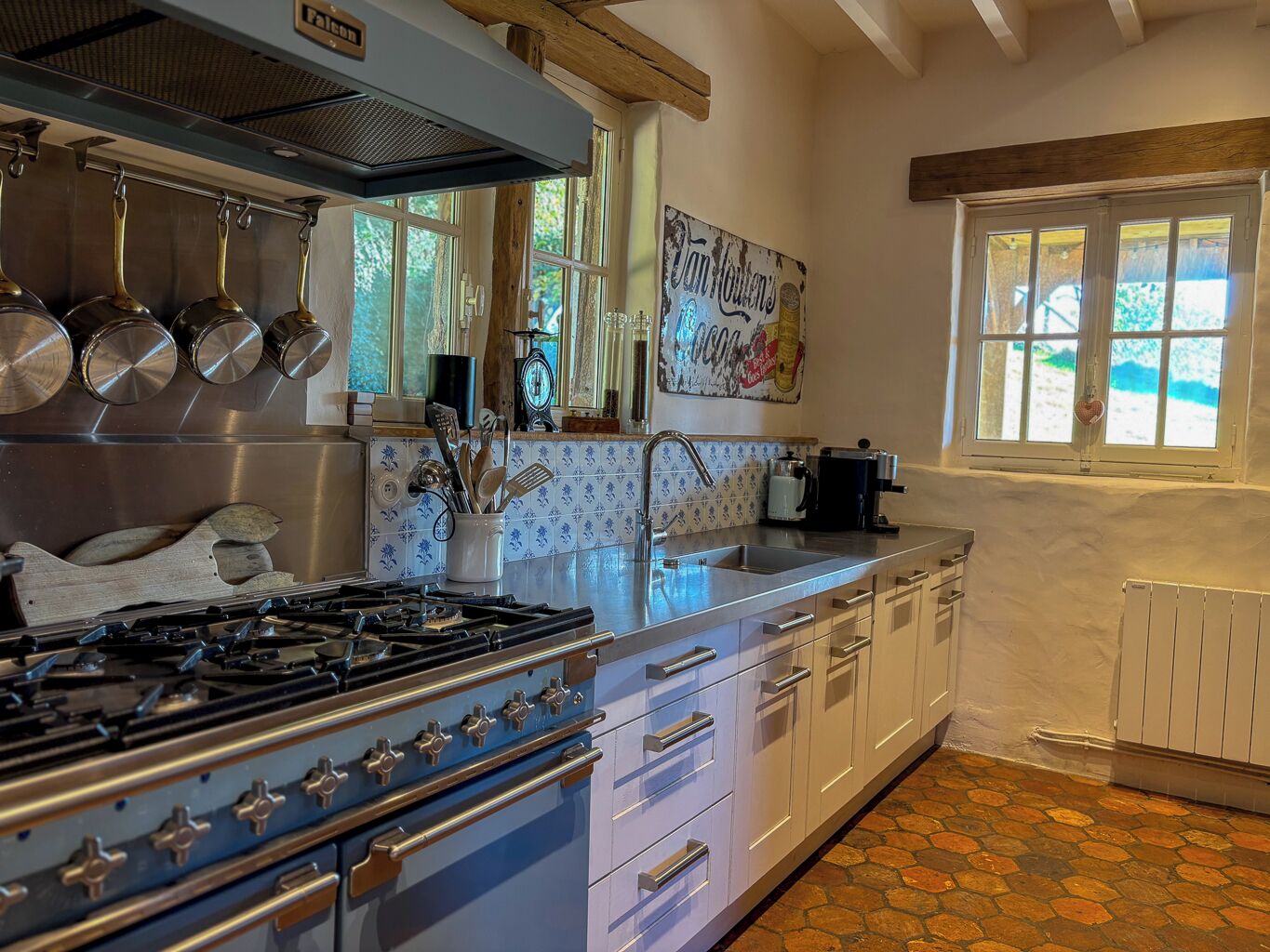 Una cocina con estufa a gas, ollas de acero inoxidable, cafetera y ventana con vista a follaje.