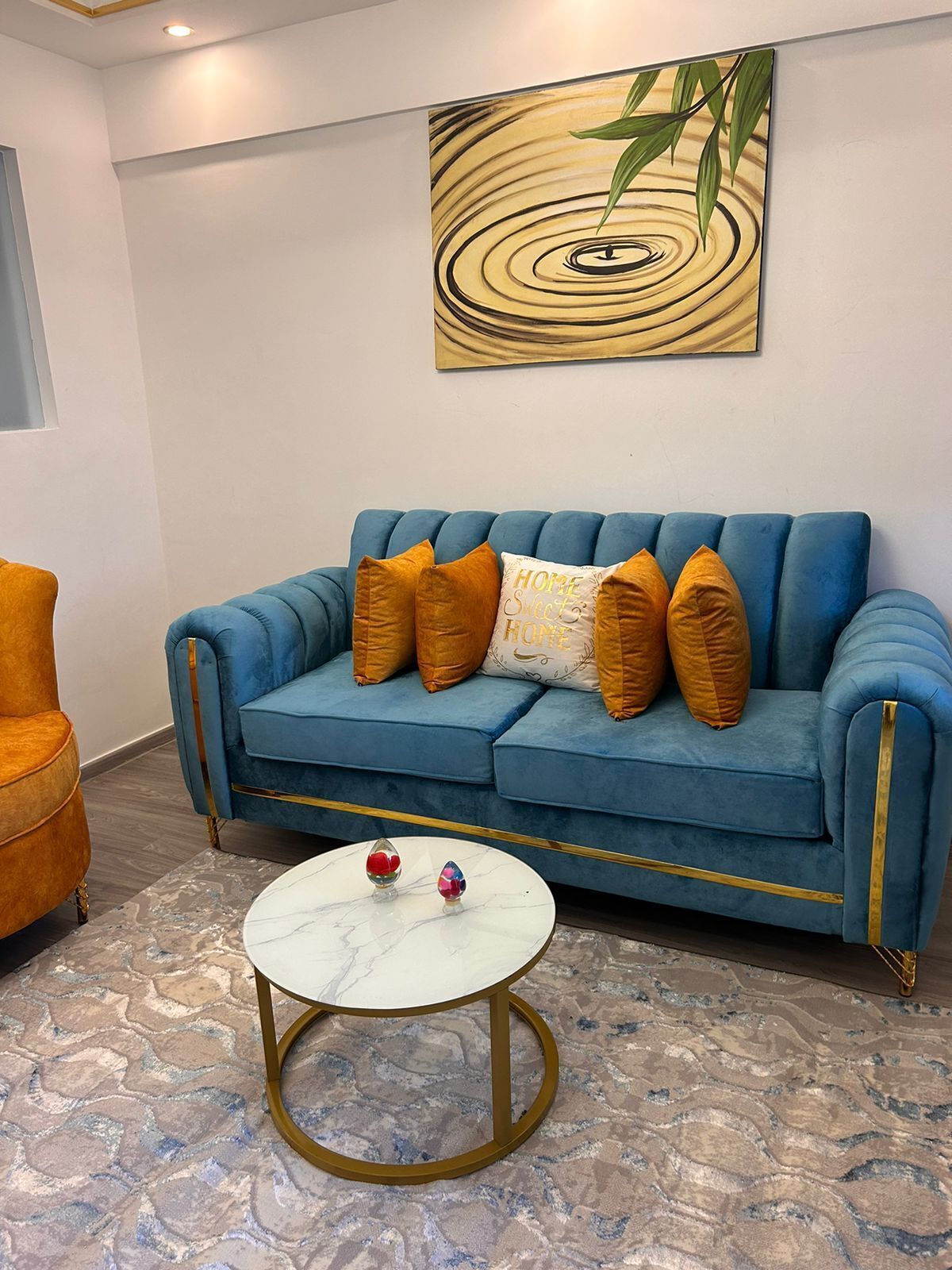 A blue sofa with gold trim and orange cushions, a white marble coffee table, and a painting of a water ripple on the wall.