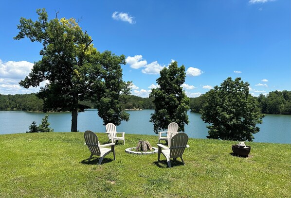 Eine Uferlandschaft mit vier Adirondack-Stühlen, die um eine Feuerstelle angeordnet sind, mit Blick auf einen stillen See und Bäume im Hintergrund.