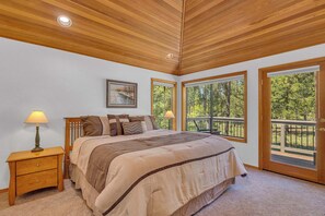 A bedroom with a large bed, wooden headboard, two bedside tables with lamps, a painting on the wall, and large windows with a view of trees. - Pet Friendly, AC, Hot tub, Ping pong, guest fav (Sunriver)