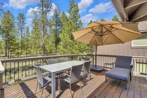 A wooden deck with a table and chairs, a large umbrella, and a view of trees. - Pet Friendly, AC, Hot tub, Ping pong, guest fav (Sunriver)