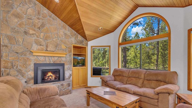 A cozy living room with a stone fireplace, leather sofa, and large windows offering a view of the outdoors. - Pet Friendly, AC, Hot tub, Ping pong, guest fav (Sunriver)