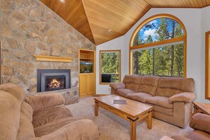 A cozy living room with a stone fireplace, leather sofa, and large windows offering a view of the outdoors. - Pet Friendly, AC, Hot tub, Ping pong, guest fav (Sunriver)
