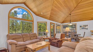A cozy living room with a high ceiling, large windows, and a dining area with wooden furniture. - Pet Friendly, AC, Hot tub, Ping pong, guest fav (Sunriver)