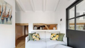 Um sofá branco com almofadas geométricas, teto com vigas de madeira, um quadro de barcos à vela e vista através de uma grande janela.