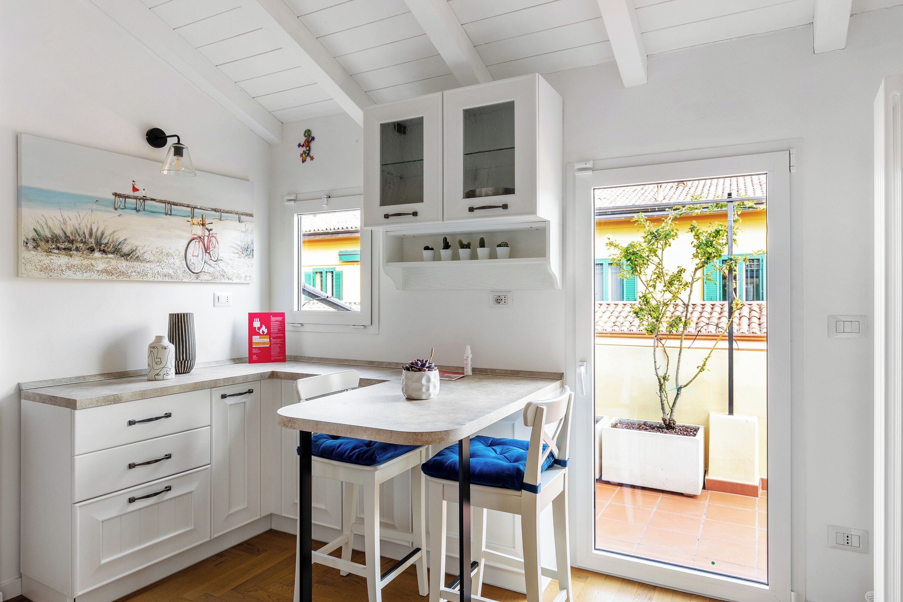 A modern kitchen with white cabinetry, a central island with blue cushions, and a view of a balcony with potted plants.