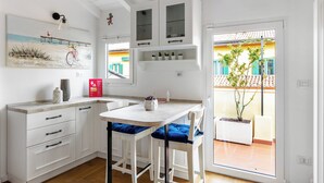 Uma cozinha moderna com armários brancos, uma ilha central com almofadas azuis e vista para uma varanda com plantas em vasos.