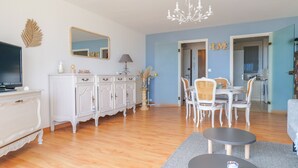 Ein Wohnzimmer mit Fernseher, weißem Schrank, Esstisch mit Stühlen und einer Deckenleuchte.
