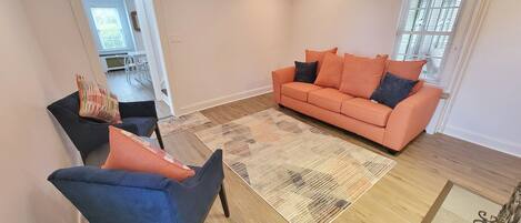 A living room with a sofa, chairs, and a coffee table. There is a window with blinds, a door leading to another room, and a patterned rug on the floor.