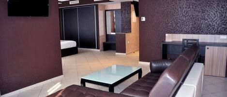 Een moderne hotelkamer met een bank, een glazen salontafel, een bed, een televisie en een kitchenette.