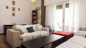 A living room with a sofa, a coffee table, a bookshelf, and a balcony with a chair. - Spacious 2 Bd Pireas Apt w Cozy Balcony (Piraeus)