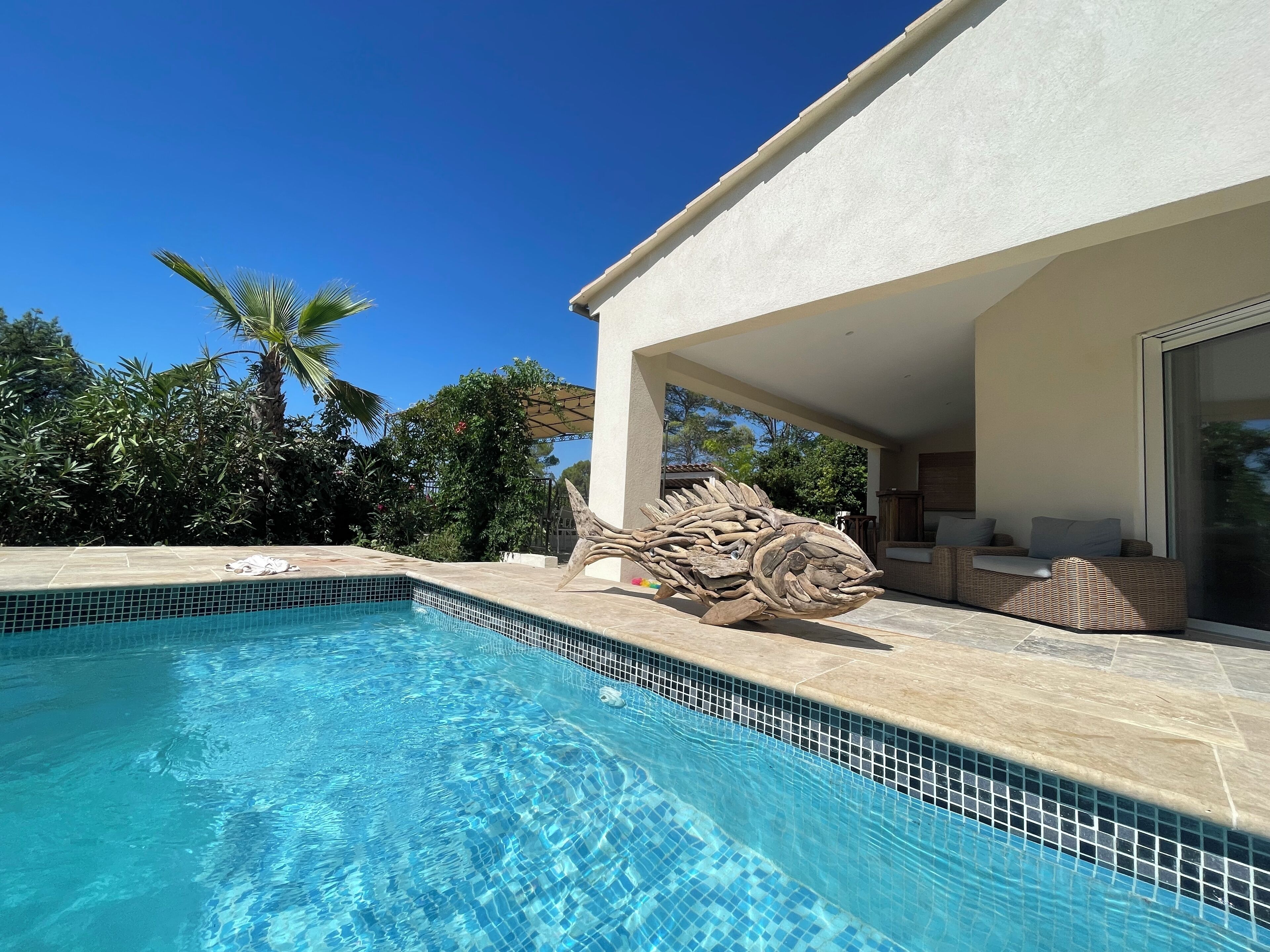 Una piscina con agua azul cristalina, una gran escultura de madera y un área de estar al aire libre techada con un sofá de mimbre.