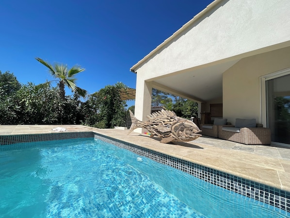 A swimming pool with clear blue water, a large wooden sculpture, and a covered outdoor seating area with a wicker sofa.