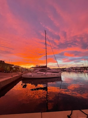 Un port de plaisance avec des bateaux amarrés, une eau calme qui reflète le ciel et un coucher de soleil aux teintes roses et orangées.