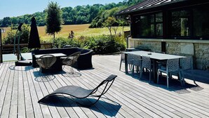 Eine Holzterrasse mit Gartenmöbeln, einem Grillbereich und einem Steinhaus mit großen Fenstern.