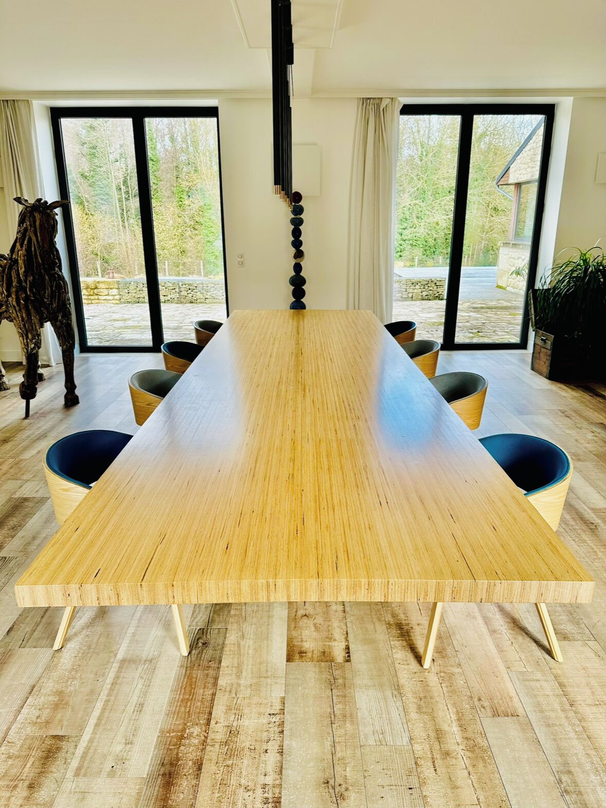 A large rectangular wooden table with four chairs, set in a room with large windows and a view of greenery outside.