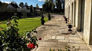 Ein gepflegter Garten umgibt einen Gebäudebereich mit Bogenfenstern, zu dem ein Steinweg führt, unter einem wolkenlosen Himmel.