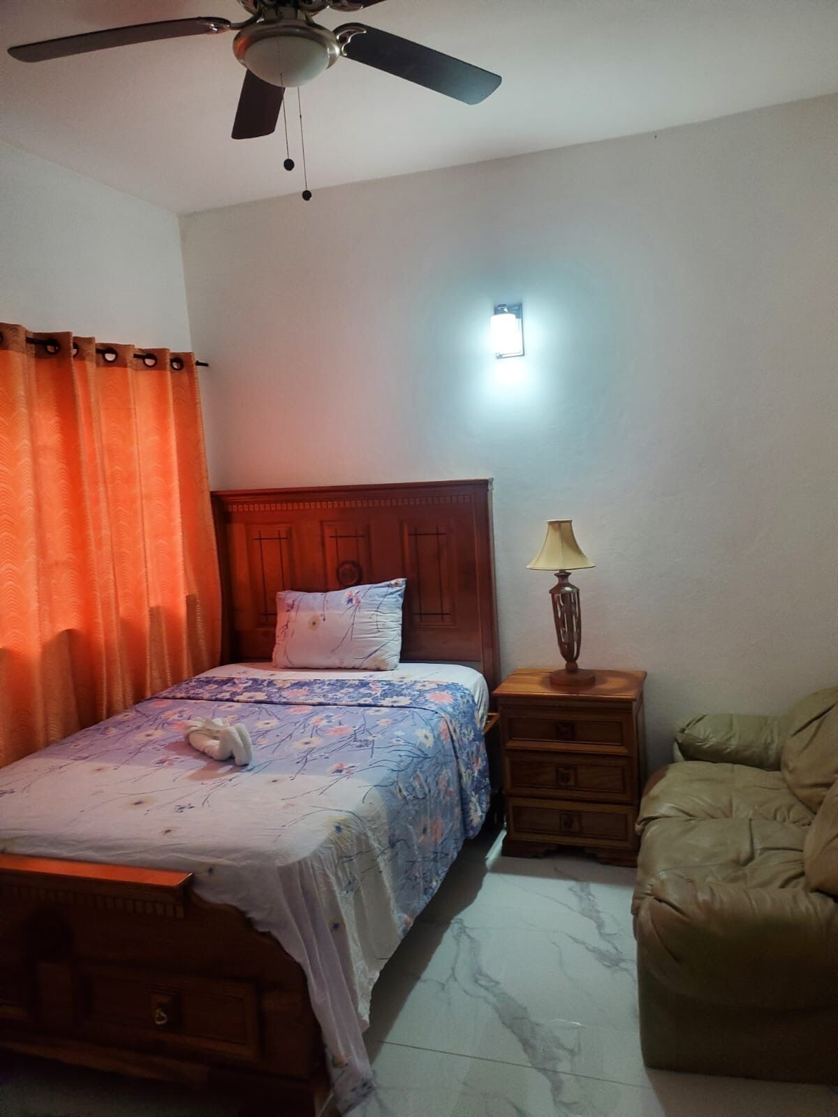 A bedroom with a bed, a nightstand, a lamp, a ceiling fan, and a sofa.