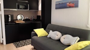 A modern living space with a grey sofa, yellow and striped pillows, a kitchen with black cabinets, a microwave, and a painting of a boat on the wall. - Studio Accès Direct Plage (Arcachon)