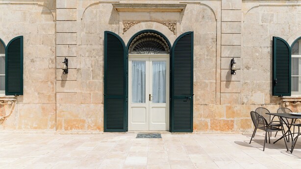 Un bâtiment doté d’une façade en pierre, d’une porte d’entrée centrale en arc surmontée de volets verts, et d’un petit espace extérieur am énagé avec des chaises.