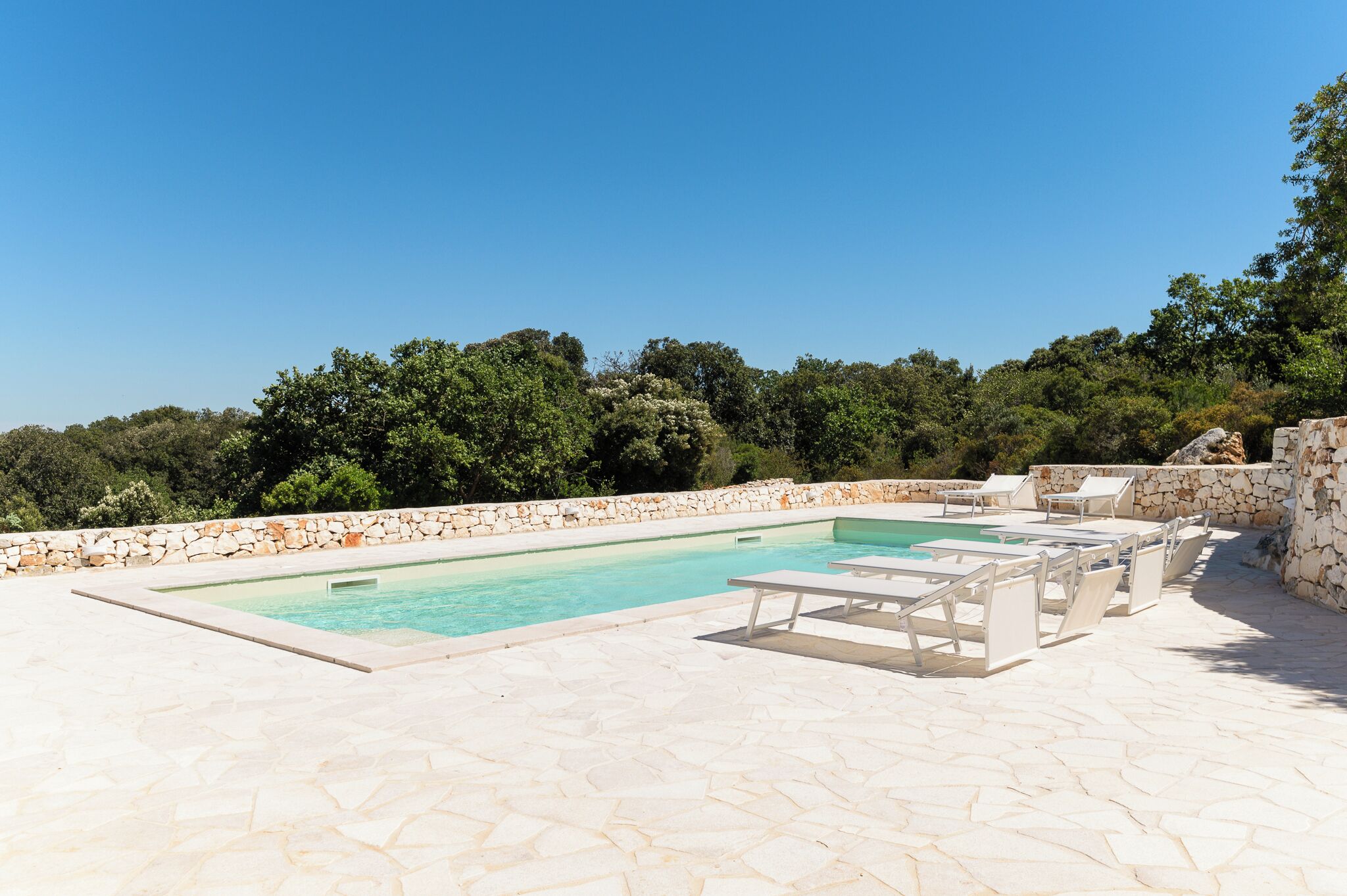 A rectangular swimming pool with a stone wall and lounge chairs.