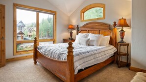 A bedroom with a large window, a wooden bed, two bedside lamps, and a view of a house and trees outside.