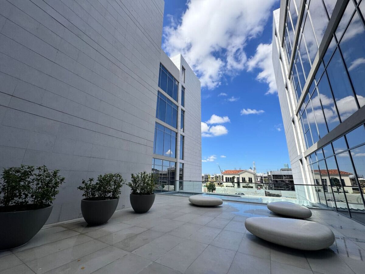 Un bâtiment moderne doté de grandes fenêtres et surplombant un ciel dégagé. La terrasse est agrémentée de plantes en pot et d’espaces aménagés pour s’asseoir.