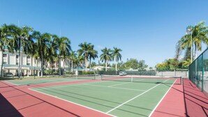 A tennis court with a red and green surface, surrounded by palm trees and a fence.