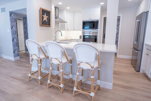 A modern kitchen with white cabinetry, a stainless steel refrigerator, and a built-in oven. There are three wicker chairs with white cushions at the island.