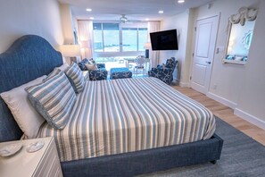 A hotel room with a large bed, a flat-screen TV, a seating area with a table and chairs, and a view of the outdoors through the windows. - This beautiful three bedroom three bathroom gulf front condo (Sarasota)