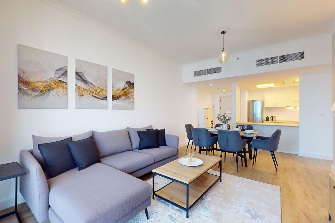 Ein modernes Wohnzimmer mit einer grauen Eckcouch, einem Holztisch und einem Essbereich mit Tisch und Stühlen. An der Wand befinden sich abstrakte Gemälde und im Hintergrund eine Küche.