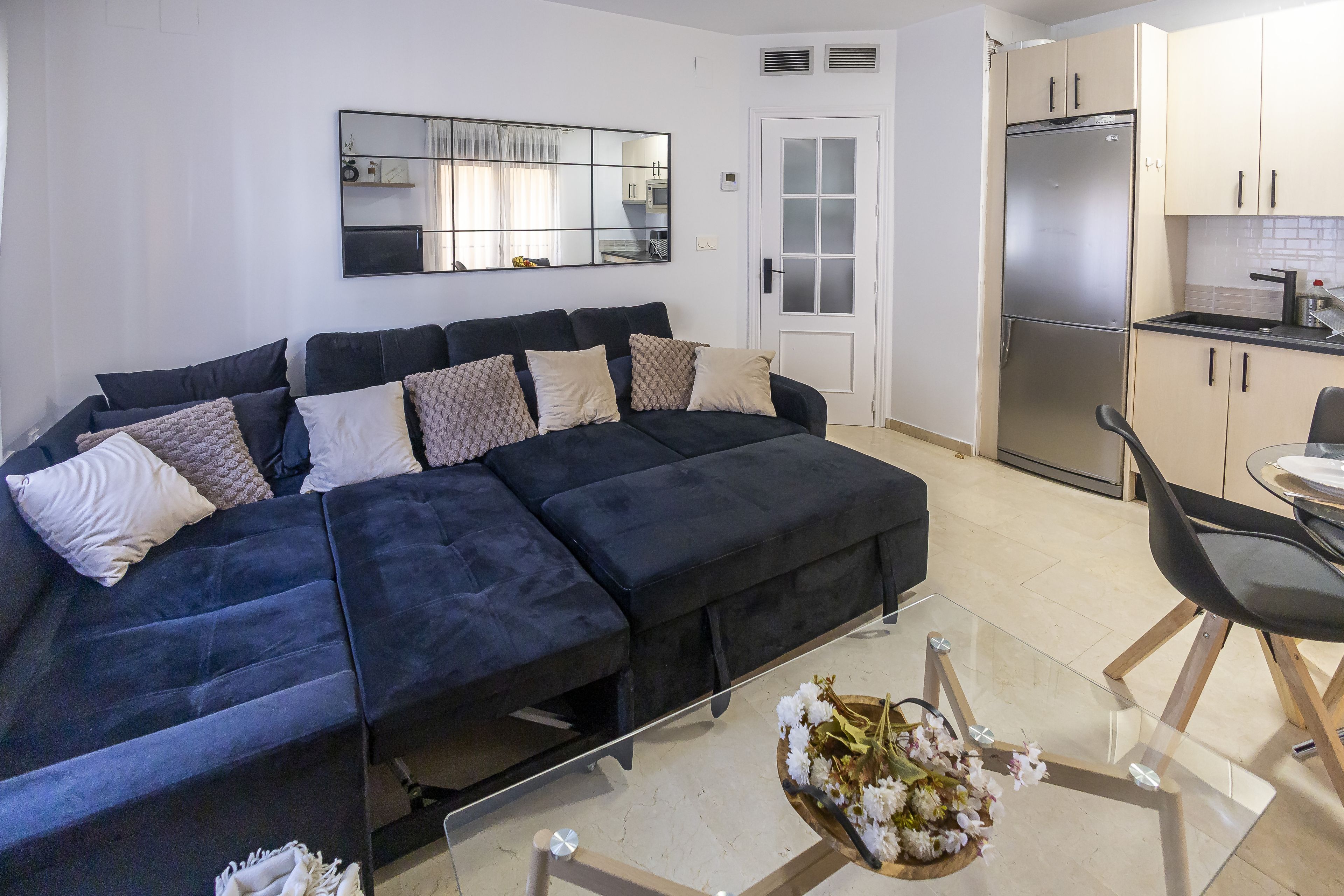 A modern living space with a dark blue sectional sofa, a glass coffee table, and a kitchen area with a stainless steel refrigerator.