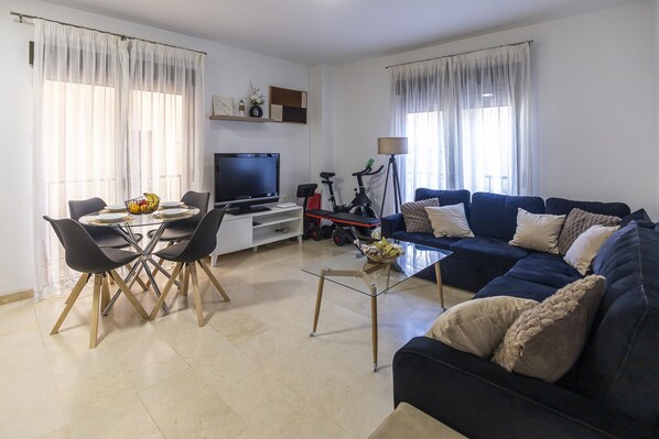 A living room with a dark blue sofa, a glass coffee table, a dining table with chairs, a television, and exercise equipment.