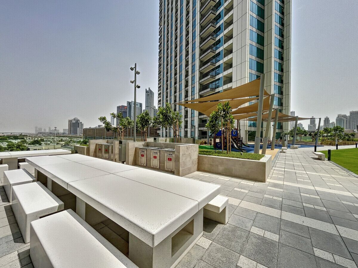 An outdoor seating area with benches and tables, surrounded by greenery and a city skyline in the background.